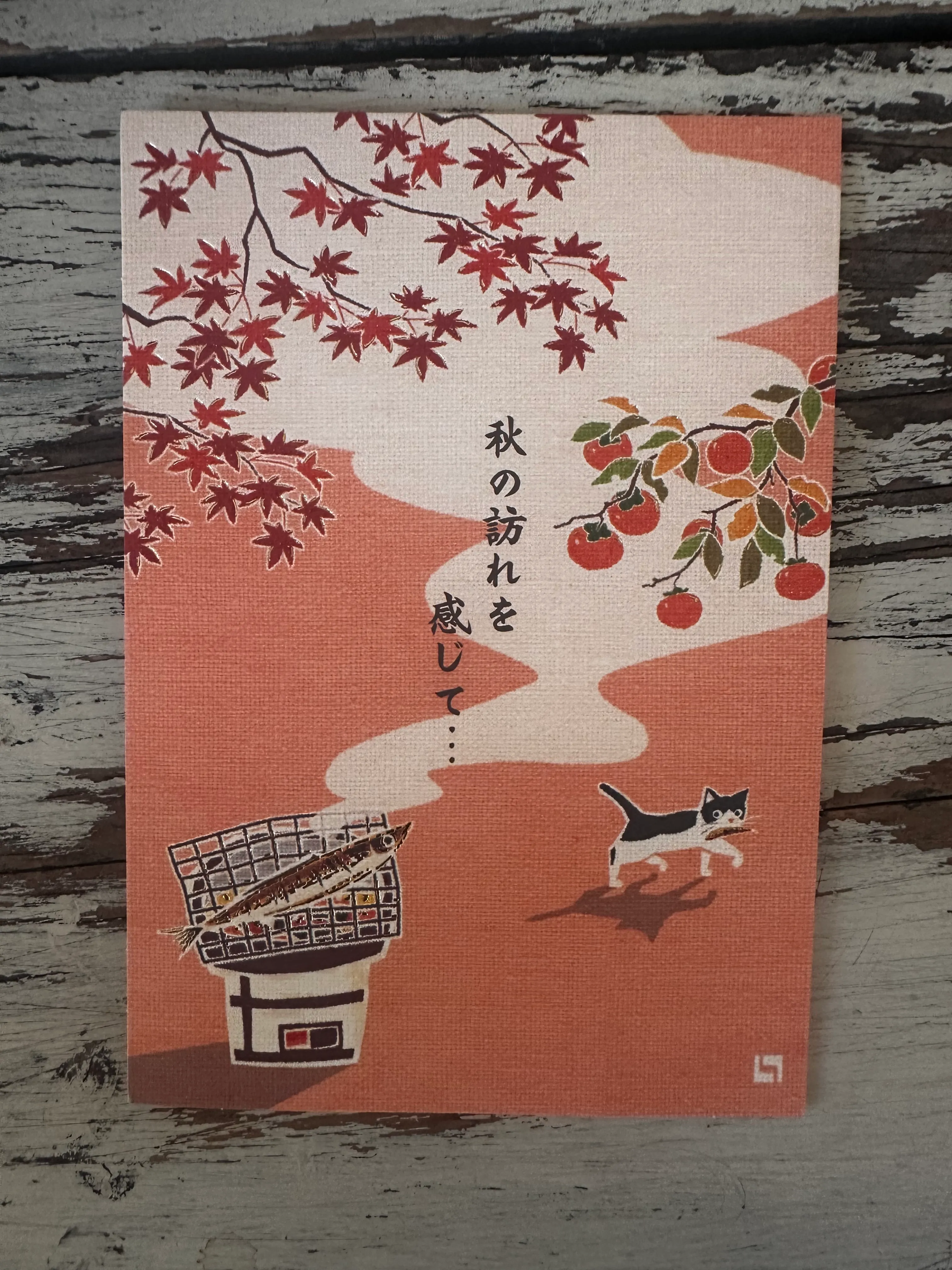 A postcard with a graphic, containing persimmons, autumn leaves, tuna being fried on a grill and a cat with fish in its mouth.