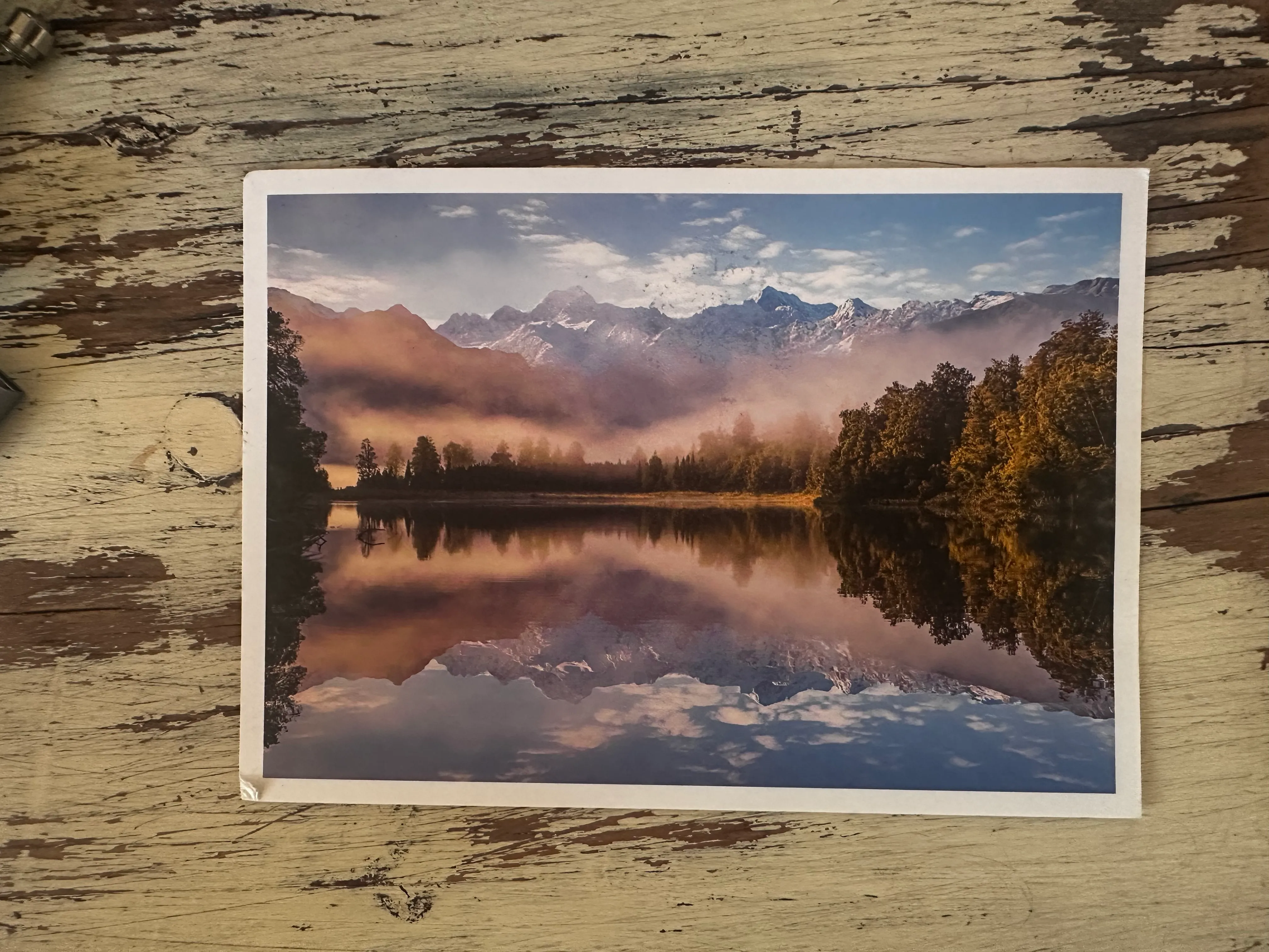 A postcard with an image on it, of a landscape with a lake in the foreground and icy mountains in the background. The lake is still and reflects the mountains behind it.
